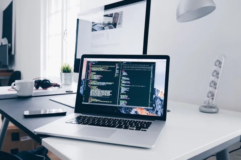 Mesa de trabalho com notebook exibindo código, monitor ao fundo e xícara de café